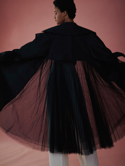 Model poses from the back with pink background in a black tulle coat with white pants under from fall 2021 collection.