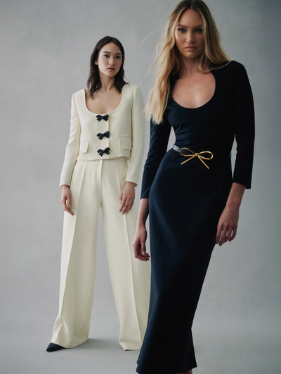 Two models pose in Fall 2021 looks. The model in the right is wearing a 3/4 sleeve black long dress with a belt around the waist and a gold bow detail in the center. The model in the left is wearing white pants with a white jacket with black button bows.