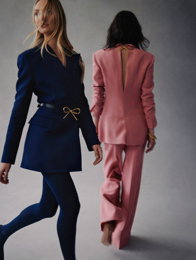 Two models in Fall 2021 Looks. Model in the left is wearing navy tights with a navy blazer and a bow belt around the waist. The model in the right is facing backwards and is wearing pink pants with a pink blazer with a cutout in the back and bow closure detail.