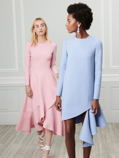 Two models pose indoors in front of a gray wall with white picture frame molding. The model on the left wears a pink long-sleeve wool dress with an asymmetrical hemline. The model on the right wears a blue long-sleeve wool shift mini dress with an asymmetrical hemline.