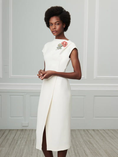 A model poses indoors in front of a gray wall with white picture frame molding, wearing an ivory sleeveless wool pencil dress with a front slit, detailed with a red flower brooch pinned to the chest.