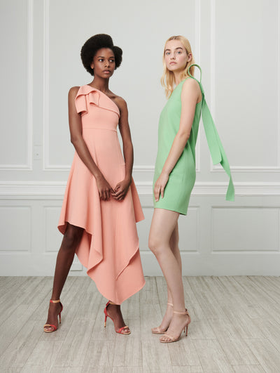 Two models pose indoors in front of a gray wall with picture frame molding. The model on the left wears an orange one-shoulder wool dress with an asymmetrical hemline. The model on the right wears a green sleeveless bow-detailed wool shift mini dress.