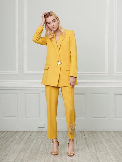 A model poses indoors in front of a gray wall with white picture frame molding. Holding a micro gold cube-like handbag, the model is wearing a yellow long-sleeve single-breasted blazer with a gold leaf button closure and yellow coordinating trousers.