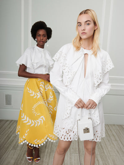 Two models pose indoors in front of a gray wall with picture frame molding. The model on the left wears a white short-sleeve ruffle-detailed blouse and a yellow skirt with white leaf embroidery and pompom trim. Holding a white cube-like handbag, the model on the right wears a white long-sleeve eyelet embroidered shift mini dress.