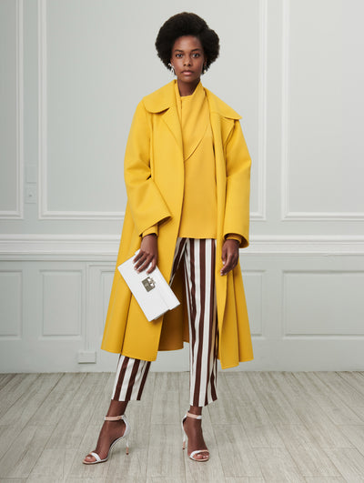A model poses indoors in front of a gray wall with white picture frame molding. Holding a white push-lock closure clutch, the model is wearing a yellow long-sleeve open-front wool coat, layered over a yellow drape-detailed long-sleeve blouse, paired with white and brown striped silk trousers.