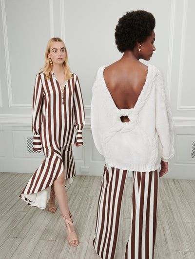 Two models pose indoors in front of a gray wall with white picture frame molding. The model on the left wears a long-sleeve V-neck white and brown striped silk dress with an asymmetrical hemline. Her back turned, the model on the right wears a white open-back knit pullover, paired with white and brown striped wide-leg silk trousers.