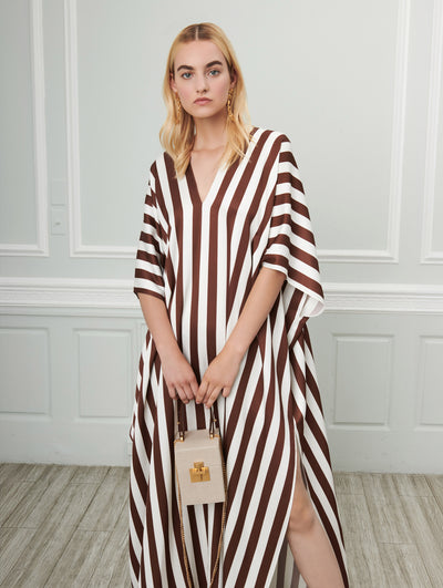 A model poses indoors in front of a gray wall with white picture frame molding. Holding a beige cube-like handbag, the model is wearing a white and brown striped V-neck silk caftan.