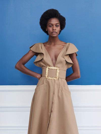 A model poses indoors in front of a blue wall with white picture frame molding, wearing a brown ruffle-detailed short-sleeve dress, accented with a statement gold leaf belt. 