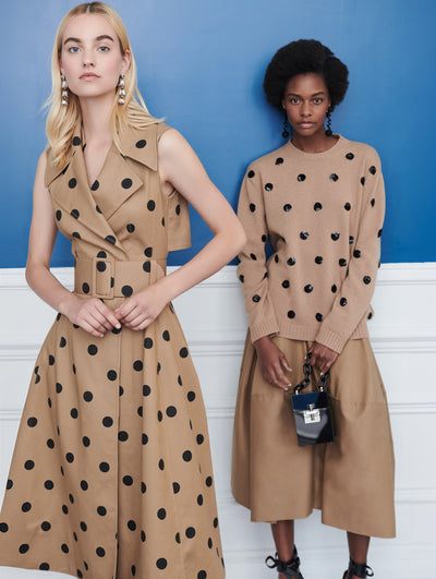 Two models pose indoors in front of a blue wall with white molding. The model on the left wears a brown trench-inspired sleeveless dress printed with black polka dots. Holding a black cube-like handbag, the model on the right wears a brown long-sleeve knit pullover printed with black polka dots and coordinating a brown skirt.