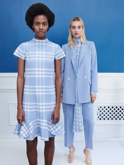 Two models pose indoors in front of a blue wall with white molding. The model on the left wears a blue and white check short-sleeve shift dress with ruffled hemline. The model on the right wears a blue long-sleeve single-breasted blazer, layer over a blue and white polka dot tie-neck long-sleeve silk blouse and coordinating blue trousers. 
