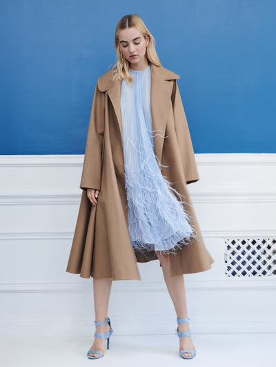A model poses indoors in front of a blue wall with white molding, wearing a brown open-front long-sleeve trench coat, layered over a blue pleated cocktail dress with feather embroidery.
