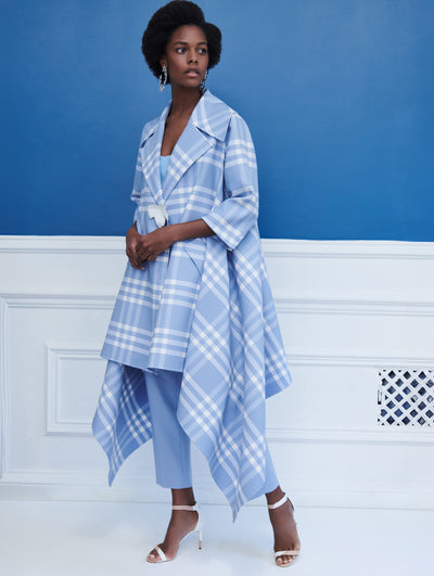 A model poses indoors in front of a blue wall with white molding, wearing a blue and white check three-quarter-length sleeve tunic with asymmetric hemline and white leaf button closure, paired with blue trousers.
