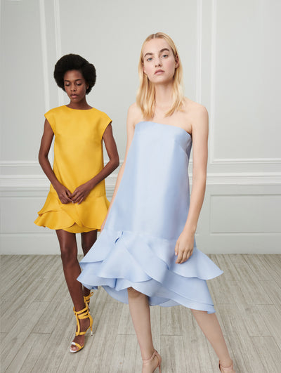 Two models pose indoors in front of a gray wall with white picture frame molding. The model on the left wears a yellow cap-sleeve shift mini dress with a ruffle hemline. The model on the right wears a blue strapless shift mini dress with a ruffle hemline.