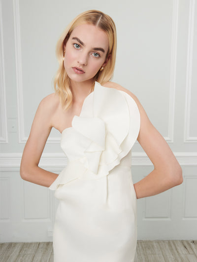 A model poses indoors in front of a gray wall with white picture frame molding, wearing a white strapless dress with a three-dimensional draped design concentrated on the bodice.