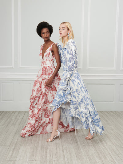 Two models pose indoors in front of a gray wall with white picture frame molding. The model on the left wears a white sleeveless V-neck maxi dress printed with a red floral toile design. The model on the right wears a white long-sleeve high-low maxi dress printed with a blue floral toile design.
