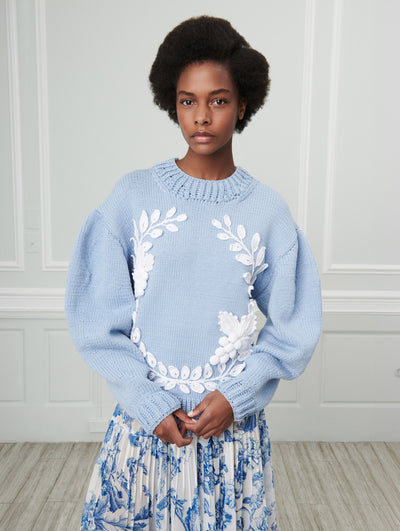 A model poses indoors in front of a gray wall with white picture frame molding, wearing a blue knit pullover with white floral embroidery and a white pleated silk skirt printed with a blue floral toile design.