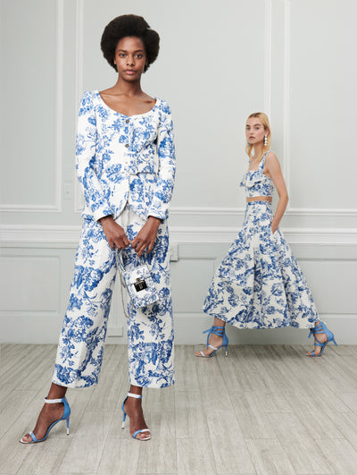Two models pose indoors in front of a gray wall with picture frame molding. Holding a white cube handbag printed with a blue floral toile design, the model on the left wears white long-sleeve scoop-neck jacket printed with a blue floral toile design and coordinating white trousers printed with a blue floral toile design. The model on the right wears a white sleeveless cropped ruffle-detail blouse printed with a blue floral toile design and a coordinating white skirt printed with a blue floral toile design.