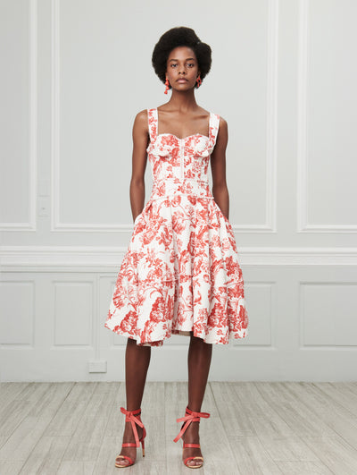 A model poses indoors in front of a gray wall with white picture frame molding, wearing a white sleeveless ruffle-detailed dress printed with a red floral toile design.