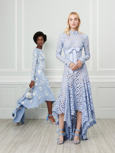 Two models pose indoors in front of a gray wall with picture frame molding. Holding a white billiards bag printed with a blue floral toile design, the model on the left wears a blue long-sleeve high-low dress with white threadwork embroidered florals. The model on the right wears a blue long-sleeve mock-neck dress with an asymmetrical hemline and a cutout floral design and bow-detailed bodice.