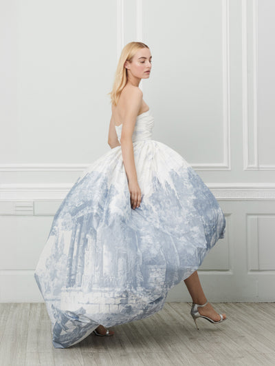A model poses indoors in front of a gray wall with white picture frame molding, wearing a white high-low evening gown printed with a blue toile design concentrated on the voluminous skirt.