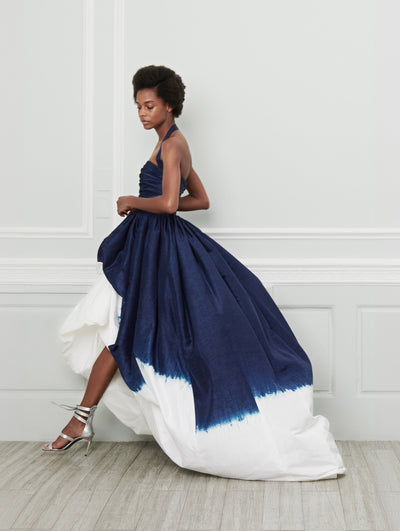 A model poses indoors in front of a gray wall with white picture frame molding, wearing an ombré halter-neck high-low evening gown where shades of navy transition to bright white.