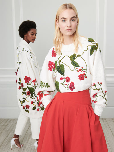 Two models pose indoors in front of a gray wall with picture frame molding. The model on the left wears a white oversized long-sleeve cardigan with multi-colored threadwork embroidered florals. The model on the right wears white cropped long-sleeve knit pullover with multi-colored threadwork embroidered florals, paired with a red skirt.