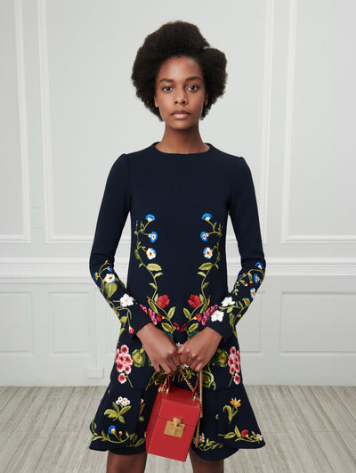 A model poses indoors in front of a gray wall with white picture frame molding. Holding a red cube-like handbag, the model is wearing a navy long-sleeve mini dress with multi-colored threadwork embroidered florals. 