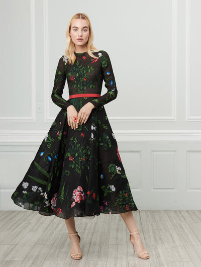 A model poses indoors in front of a gray wall with white picture frame molding, wearing a black long-sleeve fil coupé dress with a red self-belt and multi-colored threadwork embroidered florals.