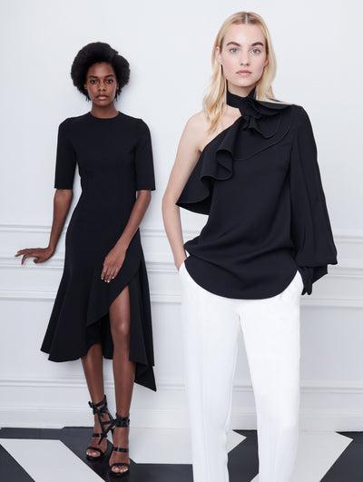Two models pose indoors in front of a gray wall with picture frame molding. The model on the left wears a black short-sleeve wool dress with an asymmetrical hemline. The model on the right wears a black one-shoulder ruffle-detailed blouse, paired with white trousers.