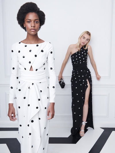 Two models pose indoors in front of a gray wall with picture frame molding. The model on the right wears a white long-sleeve evening gown printed with black polka dots. Holding a black bow-detailed clutch, the model on the right wears a black one-shoulder evening gown with a high front slit and printed with white polka dots.