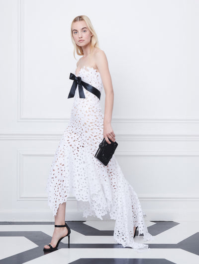 A model poses indoors in front of a gray wall with white picture frame molding. Holding a black clutch, the model is wearing a white strapless dress with an asymmetrical hemline and a cutout floral design and black bow-detailed bodice.