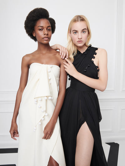 Two models pose indoors in front of a gray wall with picture frame molding. The model on the left wears a white strapless evening gown with draped detailing and pompom trim. The model on the right wears a black halter-neck evening gown with a high front slit and pompom trim.