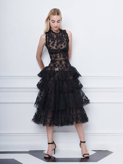 A model poses indoors in front of a white wall with picture frame molding, wearing a black sleeveless semi-sheer lace cocktail dress with scallop trim and a tiered skirt.