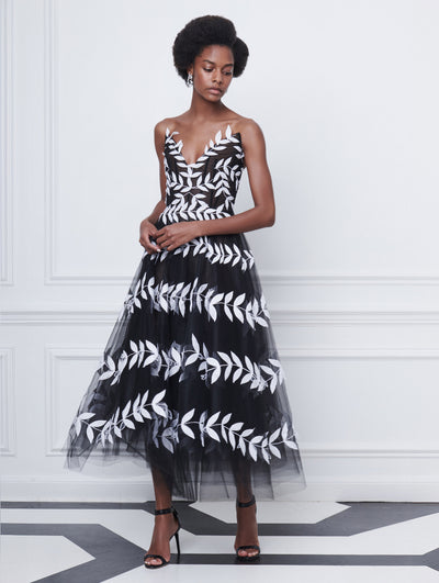 A model poses indoors in front of a white wall with picture frame molding, wearing a black strapless tulle cocktail dress with white leaf embroidery.