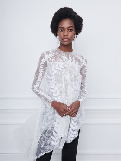 A model poses indoors in front of a white wall with picture frame molding, wearing a white long-sleeve lace blouse with leaf embroidery and an asymmetrical hemline, paired with black trousers.