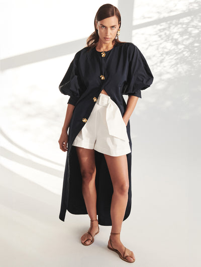 A model wears white shorts and a long black blouse with gold button flowers