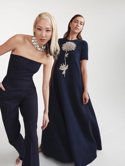 Two models are shown; the model on the left wears a black pantsuit with a big, jeweled necklace and the model on the right wears a long black dress with an embroidered silver flower