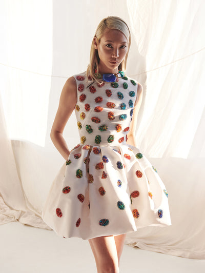 The model wears a short white dress with rainbow dot details and a rainbow collar