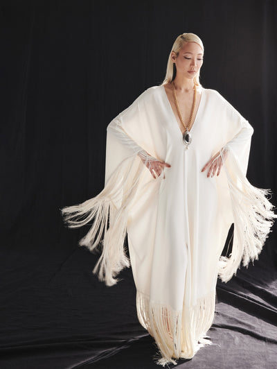 A model wears a long white dress with fringe on the bottom and sleeves