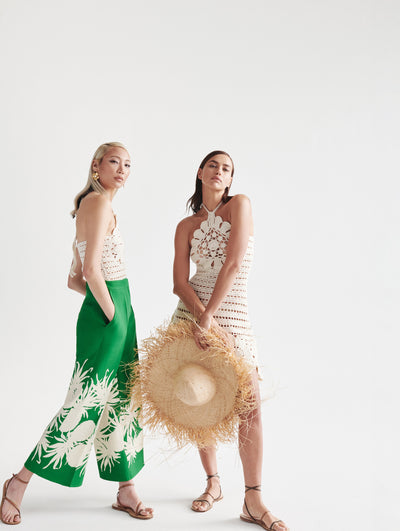 Two models; the model on the left wears green pants with pineapples embroidered in white and a crochet top and the model on the right wears a crochet white dress