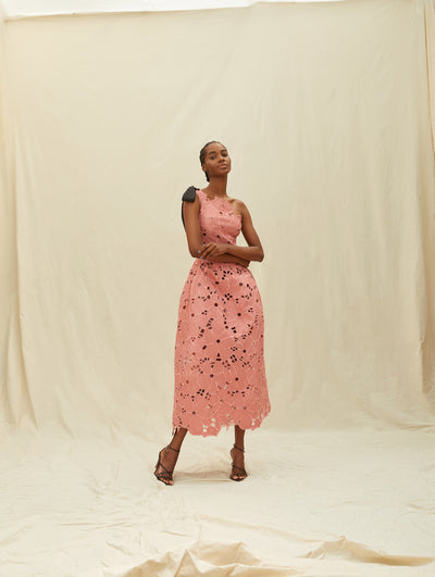 A model wears a pink lace one shoulder dress with a black bow on the shoulder