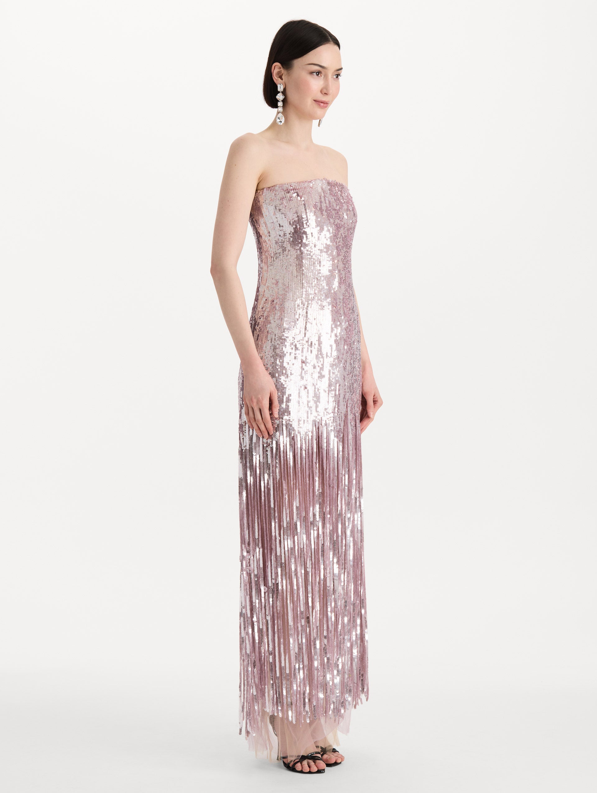 The model is wearing the Sequin Fringe Strapless Gown in the color tulip pink featuring a strapless silhouette, all-over sequins, and fringe skirt. Side View.
