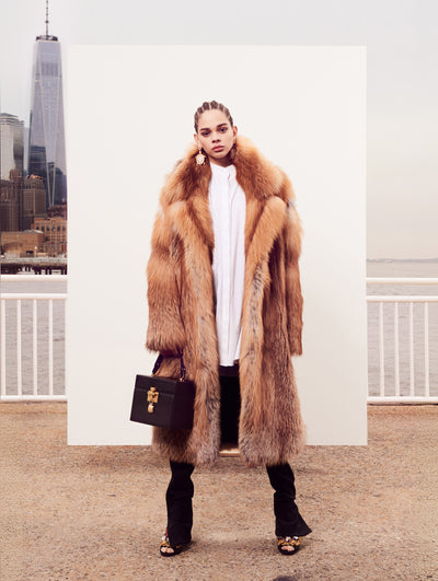 The model poses in front of a white backdrop with the New York City skyline behind her. Holding a black cube-like handbag, the model is wearing a brown open-front fur coat layered over a white button-up blouse and black trousers.