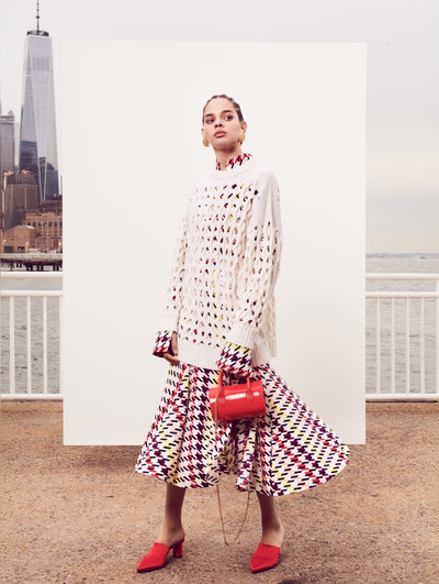 The model poses in front of a white backdrop with the New York City skyline behind her. Holding a red tube-like handbag, the model is wearing a silk long-sleeve dress printed with a multi-colored checked design layered over a white cable knit pullover.