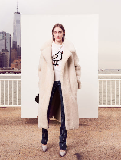 The model poses in front of a white backdrop with the New York City skyline behind her, wearing an open-front cream-colored fur coat layered over a white spread-collar pullover embroidered with a sequin bird and navy and white pinstripe trousers.