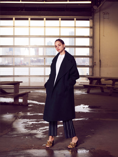 The model poses in a warehouse with a garage door behind her, revealing the New York City skyline. Two wooden picnic tables are visible in the background as she wears an oversized open-front long-sleeve navy wool coat layered over a white button-up blouse and navy and white pinstripe trousers.