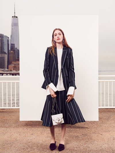 The model poses in front of a white backdrop with the New York City skyline behind her. Holding a white gardenia appliqué handbag, the model is wearing a navy and white pinstripe long-sleeve single-breasted blazer layered over a white long-sleeve blouse and a coordinating navy and white pinstripe skirt.