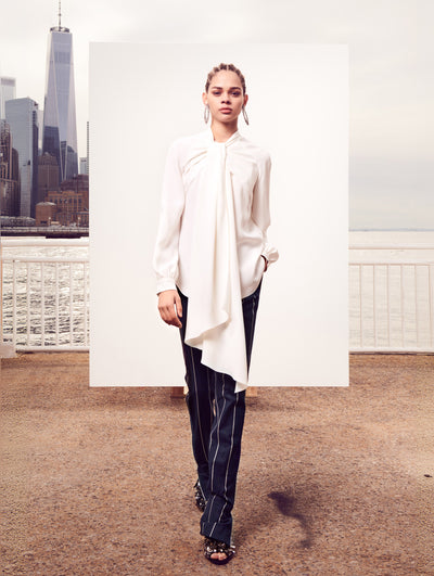 The model poses in front of a white backdrop with the New York City skyline behind her, wearing a white long-sleeve blouse with a dramatic self-tie neck sash and navy and white pinstripe trousers.