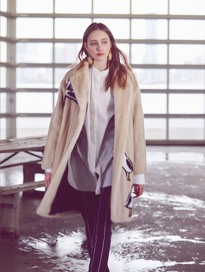 The model poses in a warehouse with a garage door behind her, revealing the New York City skyline. A single wooden picnic table is visible in the background as she wears an oversized cream-colored fur coat detailed with birds layered over a white button-up blouse and navy and white pinstripe trousers.