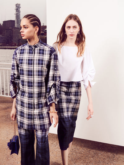 Two models pose in front of a white backdrop with the New York City skyline behind them. Holding a blue gardenia appliqué handbag, the model on the left is wearing a navy and white plaid button-up blouse and coordinating navy and white plaid trousers. The model on the right is wearing a white three-quarter-length sleeve blouse and a navy and white plaid skirt.